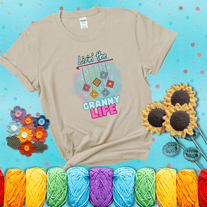 Colorful crochet granny squares hang from a hook with text “Livin’ the Granny Life” in bold pink and blue font, a fun nod to classic crochet showing a beige tee with a teal sunflower background