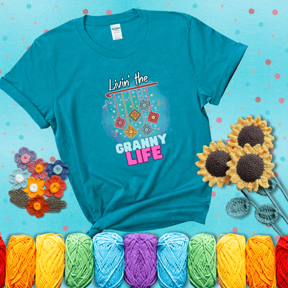 Colorful crochet granny squares hang from a hook with text “Livin’ the Granny Life” in bold pink and blue font, a fun nod to classic crochet showing a blue-green tee with a teal sunflower background