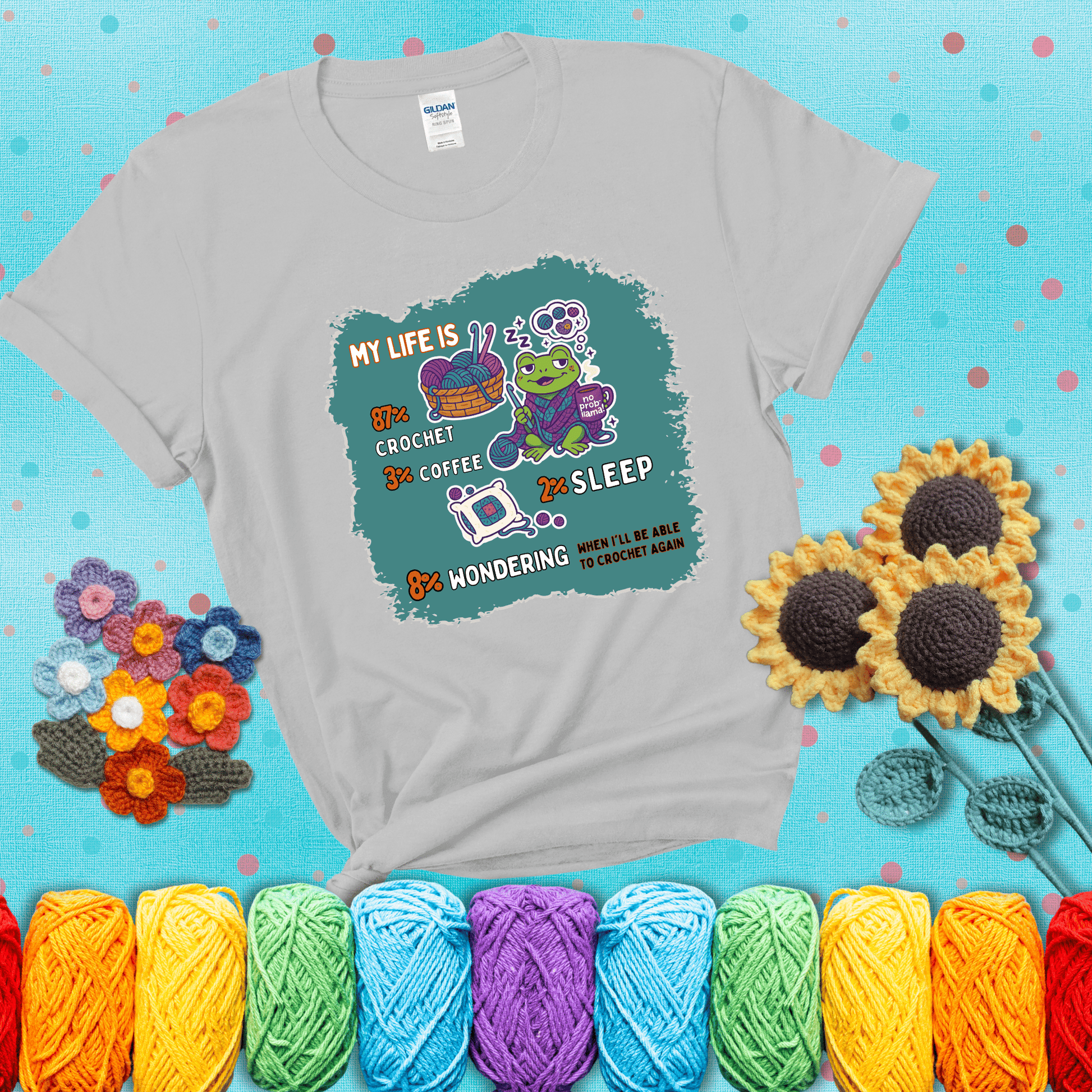 A relaxed frog crocheting while sipping coffee, surrounded by yarn and dreaming of more yarn. Includes whimsical text dividing life into percentages: 87% crochet, 3% coffee, 2% sleep, 8% wondering when to crochet again showing a light grey tee with a teal sunflower background