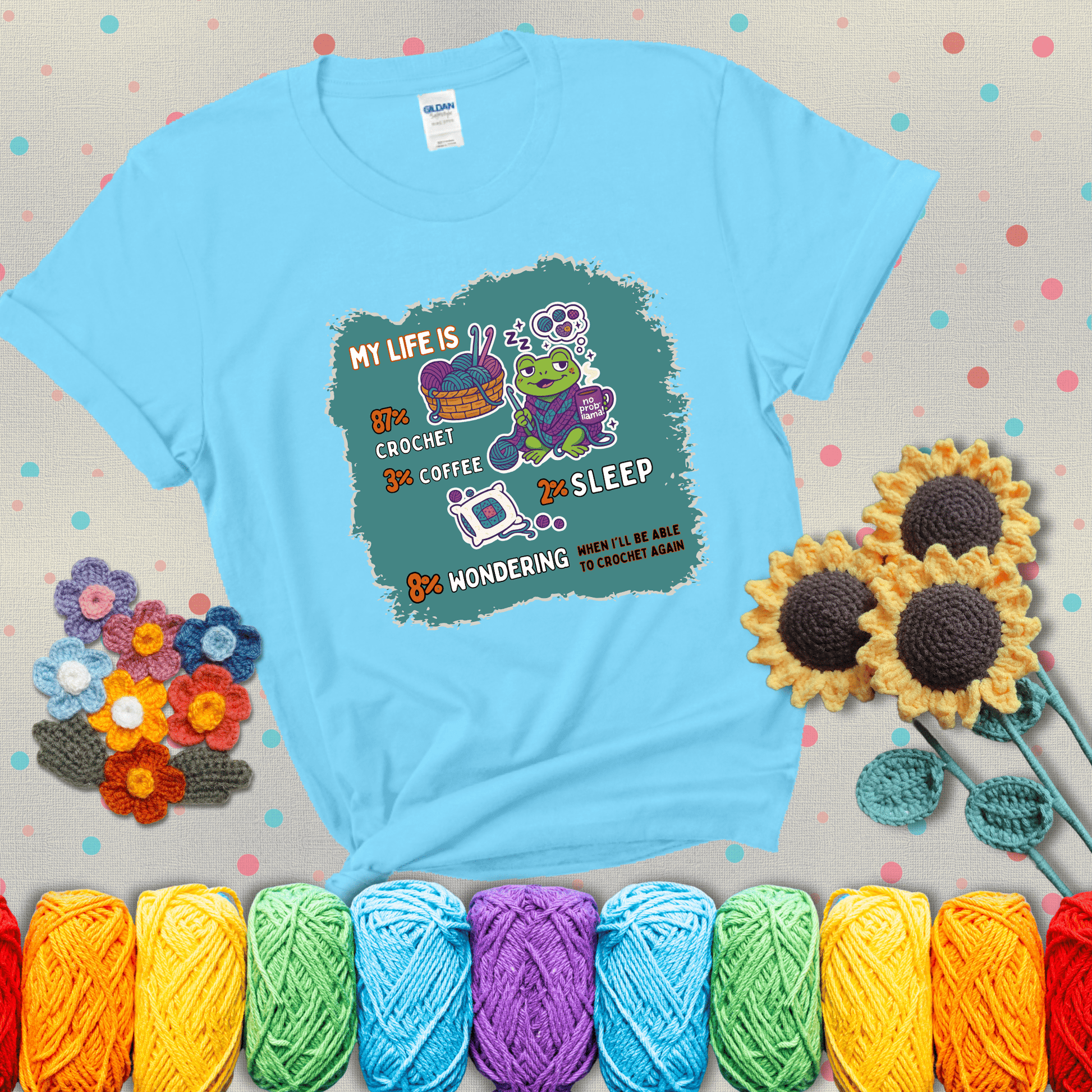 A relaxed frog crocheting while sipping coffee, surrounded by yarn and dreaming of more yarn. Includes whimsical text dividing life into percentages: 87% crochet, 3% coffee, 2% sleep, 8% wondering when to crochet again showing a light blue tee with grey sunflower background