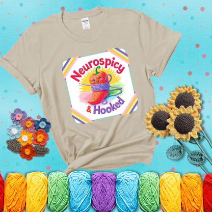 A cheerful red pepper wrapped in purple yarn with a crochet hook, framed by pastel rainbow corners and the words "Neurospicy & Hooked" showing a beige tee with a teal sunflower background