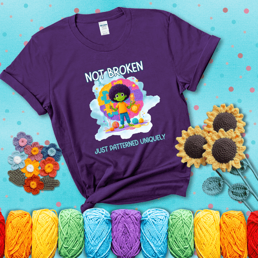 A smiling green character stands on a colorful crochet mat shaped like puzzle pieces, holding yarn and a hook, with the words “Not broken, just patterned uniquely” showing a purple tee with a teal sunflower background