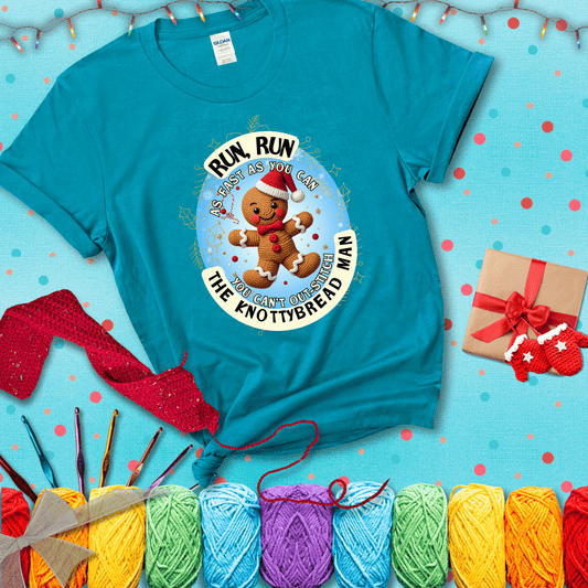 Cute crocheted gingerbread man in Santa hat with playful text: “Run, Run As Fast As You Can, You Can’t Out-Stitch the Knottybread Man.” A festive crochet pun shirt, showing a blue-green tee with a festive Christmas background