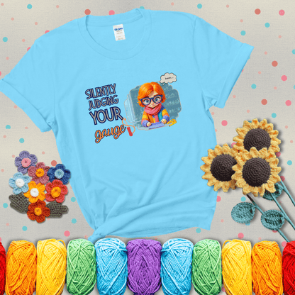 A serious-looking crocheter with rainbow clothes, big glasses, and a tape measure silently examines their stitches under the phrase “Silently judging your gauge” showing a light blue tee with grey sunflower background