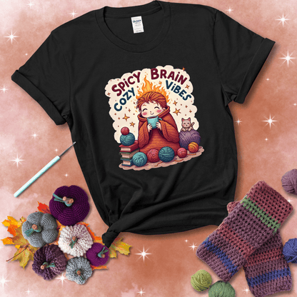 A person with flaming red hair wrapped in a chunky crochet blanket, holding a mug, surrounded by yarn balls, books, a cat, and the phrase "Spicy Brain Cozy Vibes" showing a black tee with a festive Fall background