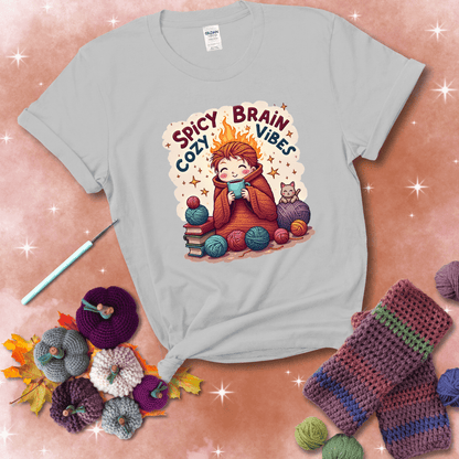 A person with flaming red hair wrapped in a chunky crochet blanket, holding a mug, surrounded by yarn balls, books, a cat, and the phrase "Spicy Brain Cozy Vibes" showing a light grey tee with a festive Fall background