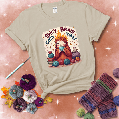 A person with flaming red hair wrapped in a chunky crochet blanket, holding a mug, surrounded by yarn balls, books, a cat, and the phrase "Spicy Brain Cozy Vibes" showing a beige tee with a festive Fall background