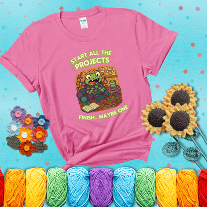 A wide-eyed crochet frog sits tangled in yarn with dozens of projects around, surrounded by chaos and “new project” notes. Caption reads “Start all the projects, finish... maybe one” showing a pink tee with a teal sunflower background