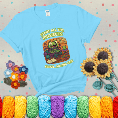 A wide-eyed crochet frog sits tangled in yarn with dozens of projects around, surrounded by chaos and “new project” notes. Caption reads “Start all the projects, finish... maybe one” showing a light blue tee with grey sunflower background