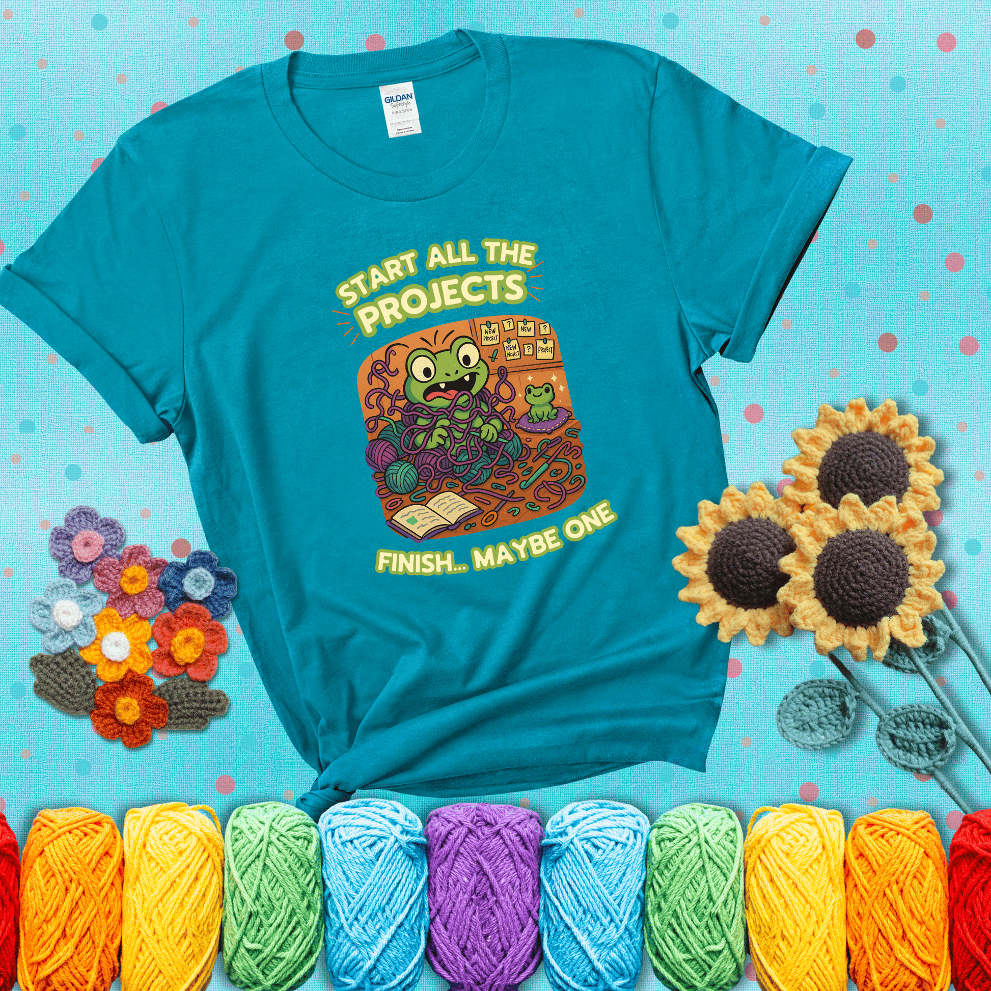 A wide-eyed crochet frog sits tangled in yarn with dozens of projects around, surrounded by chaos and “new project” notes. Caption reads “Start all the projects, finish... maybe one” showing a blue-green tee with a teal sunflower background