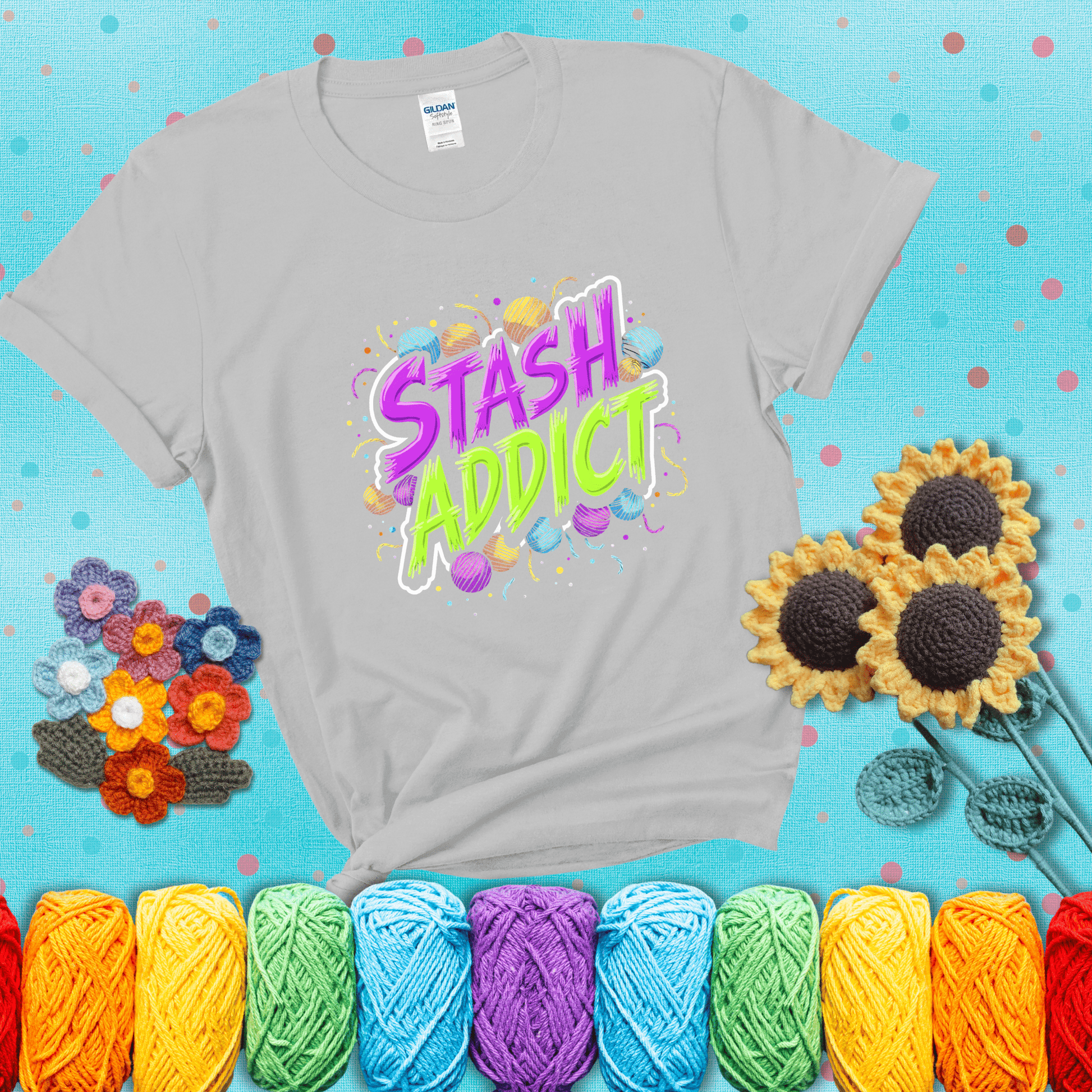 Bold neon text reading "Stash Addict" surrounded by colorful yarn balls and festive confetti on a neutral background showing a light grey tee with a teal sunflower background