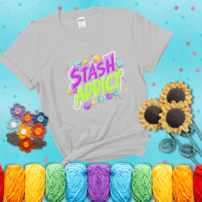 Bold neon text reading "Stash Addict" surrounded by colorful yarn balls and festive confetti on a neutral background showing a light grey tee with a teal sunflower background