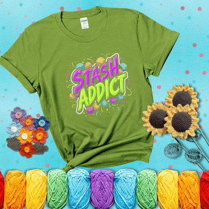 Bold neon text reading "Stash Addict" surrounded by colorful yarn balls and festive confetti on a neutral background showing a bright green tee with a teal sunflower background