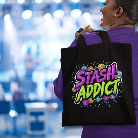 Person holding a black ‘Stash Addict’ crochet-themed tote bag with colorful yarn graphics, in an indoor stage setting.