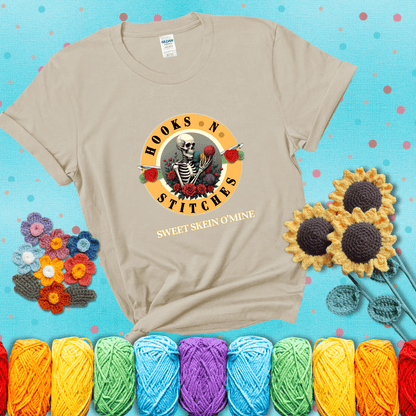 A skeleton holding colorful yarn, surrounded by roses, framed in a circular logo-style design that says “Hooks 'N Stitches” and “Sweet Skein O’Mine,” with crochet hooks piercing through showing a beige tee with a teal sunflower background