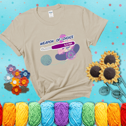 Illustration of a crocheter sitting cross-legged with a 9.0 mm crochet hook above her, labeled “Weapon of Choice.” She’s crocheting a colorful mandala-style project from a large yarn ball, showing a beige tee with a teal sunflower background