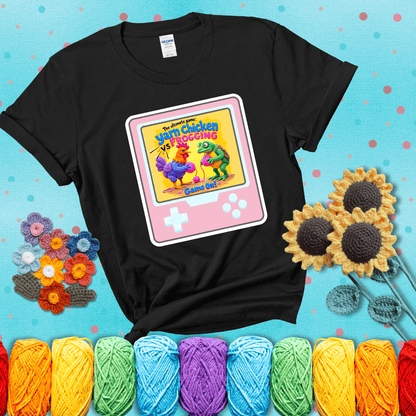 A colorful chicken and frog holding pink yarn face off inside a retro handheld game screen with the title “The ultimate game: Yarn Chicken vs. Frogging – Game On!” showing a black tee with a teal sunflower background