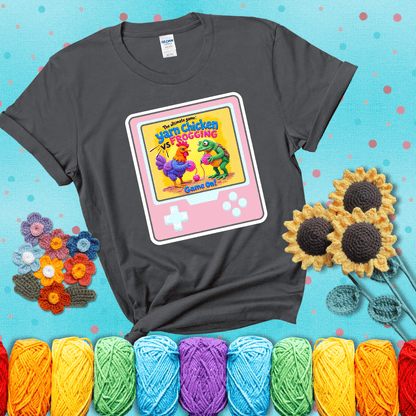 A colorful chicken and frog holding pink yarn face off inside a retro handheld game screen with the title “The ultimate game: Yarn Chicken vs. Frogging – Game On!” showing a dark grey tee with a teal sunflower background