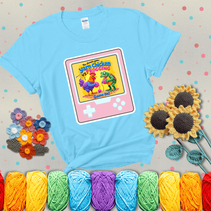A colorful chicken and frog holding pink yarn face off inside a retro handheld game screen with the title “The ultimate game: Yarn Chicken vs. Frogging – Game On!” showing a light blue tee with grey sunflower background