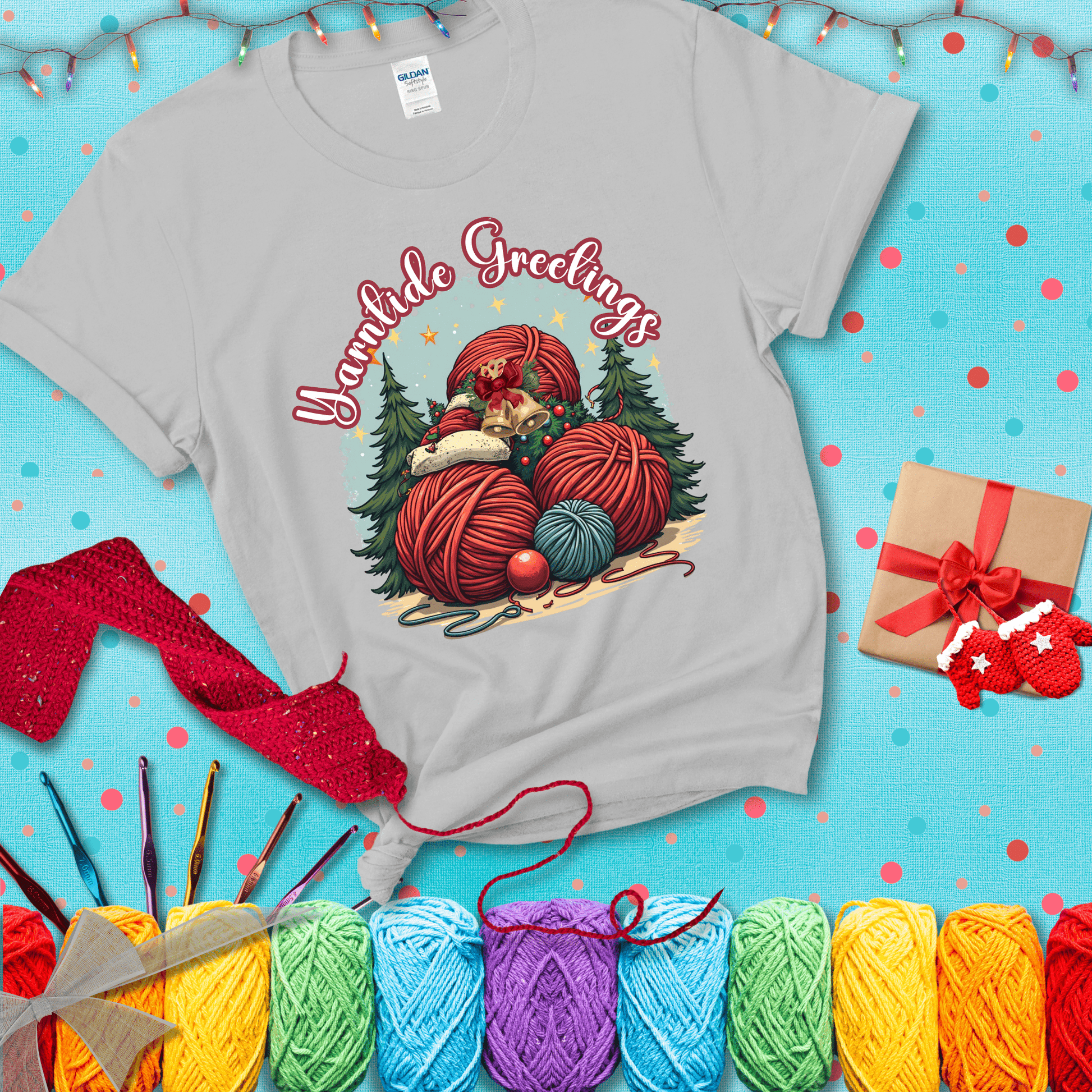 Pile of colorful yarn balls with bells, bows, and trees, with the phrase “Yarntide Greetings.” Cozy and festive holiday crochet tee, showing a light grey tee with a festive Christmas background