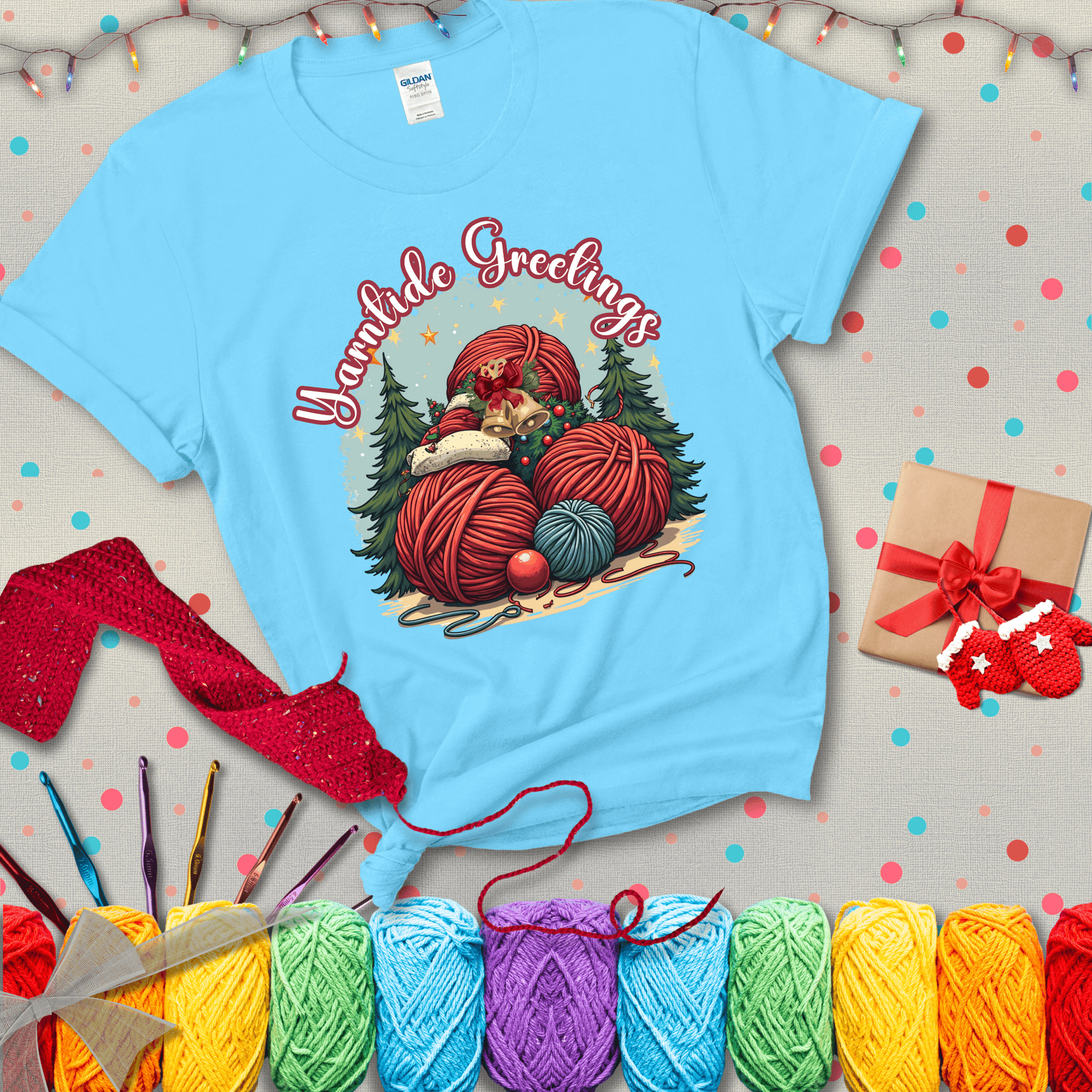 Pile of colorful yarn balls with bells, bows, and trees, with the phrase “Yarntide Greetings.” Cozy and festive holiday crochet tee, showing a light blue tee with a festive Christmas background